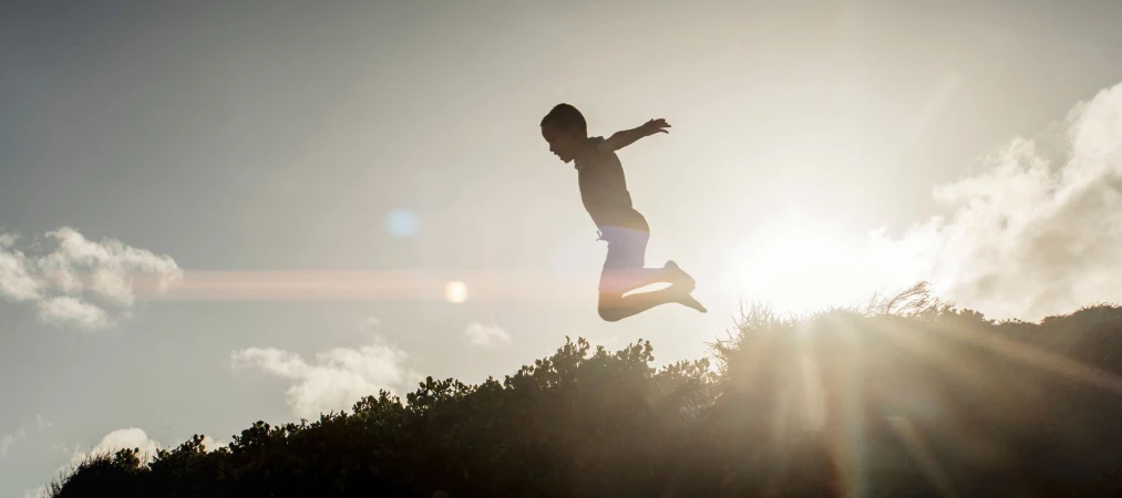 Silhouette of a child jumping mid-air against a bright sunset sky and silhouetted foliage, photographed by Dick Sweeney.