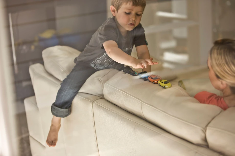 A young child is playing with toy cars on a white leather couch, indoors, with a woman sitting nearby, viewed through a window.