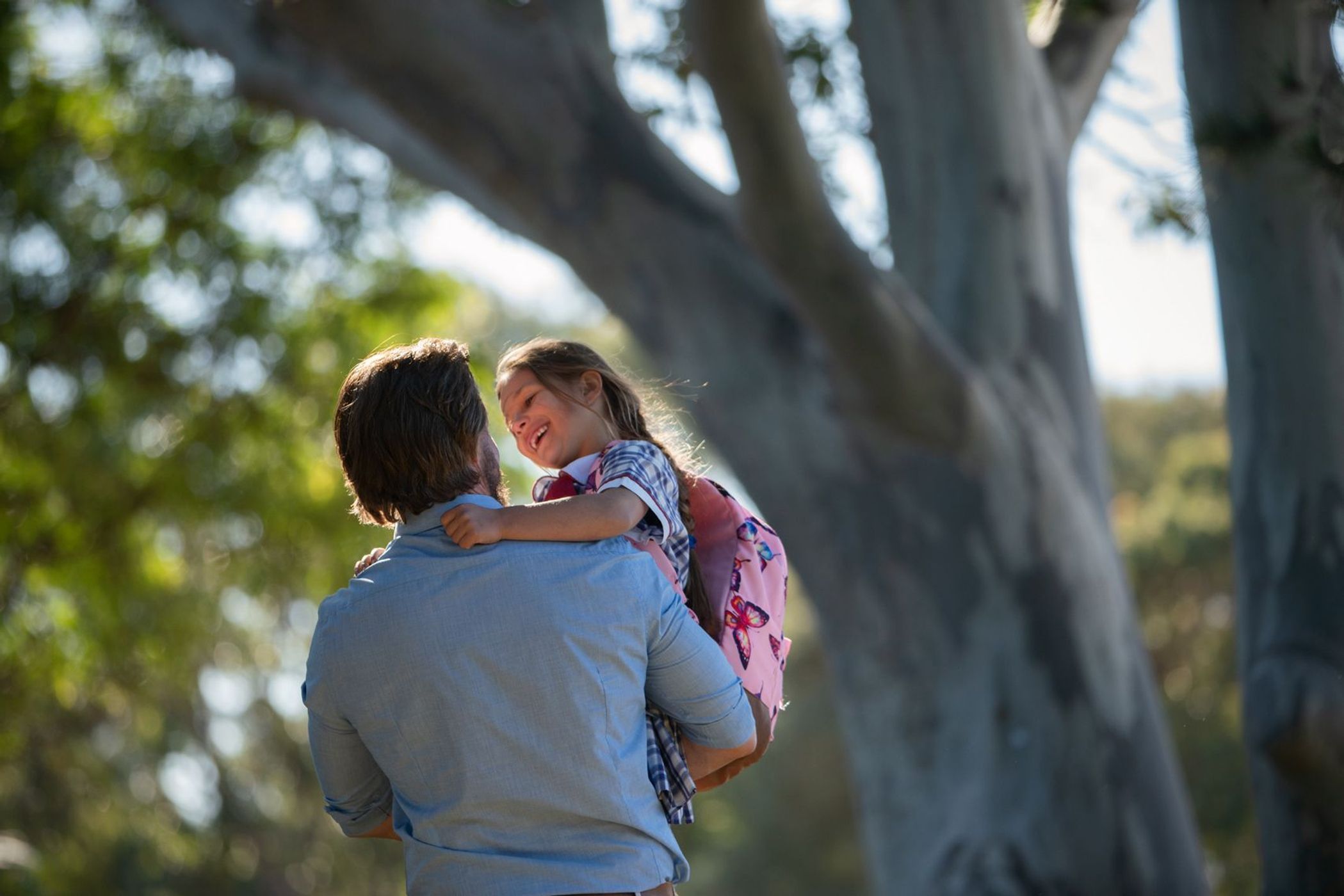 A father holds his smiling daughter in a leafy outdoor setting. The young girl has her arms around his neck. He is holding her close.