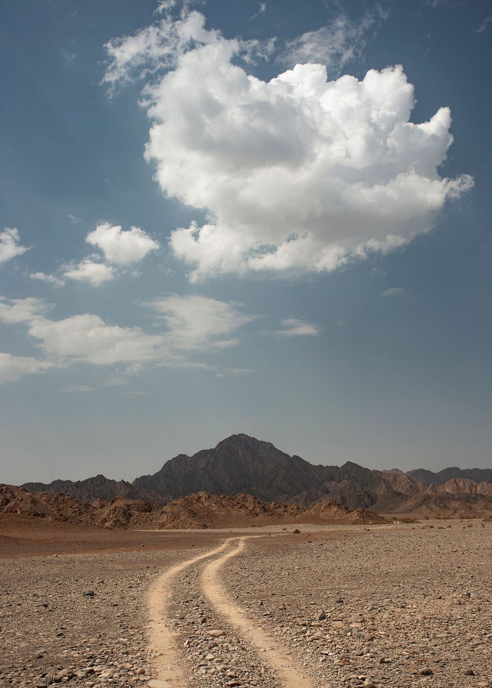 Dirt road leading to a mountain range under a blue sky with scattered clouds, creating a vast and open desert landscape.
