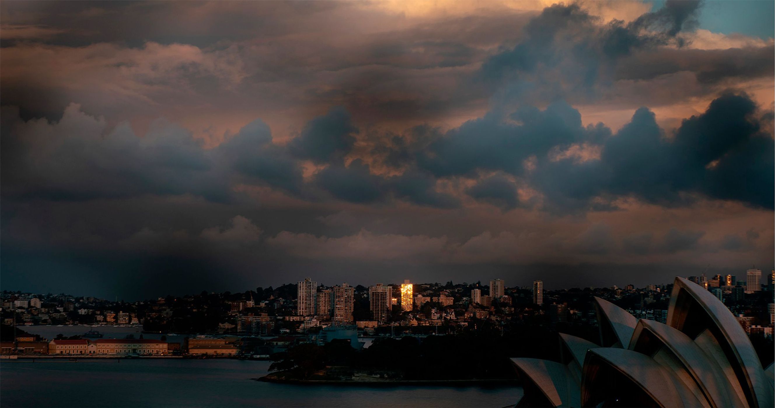 Panoramic cityscape view of the Sydney Opera House and skyline at dusk, featuring dramatic clouds and the waterfront.