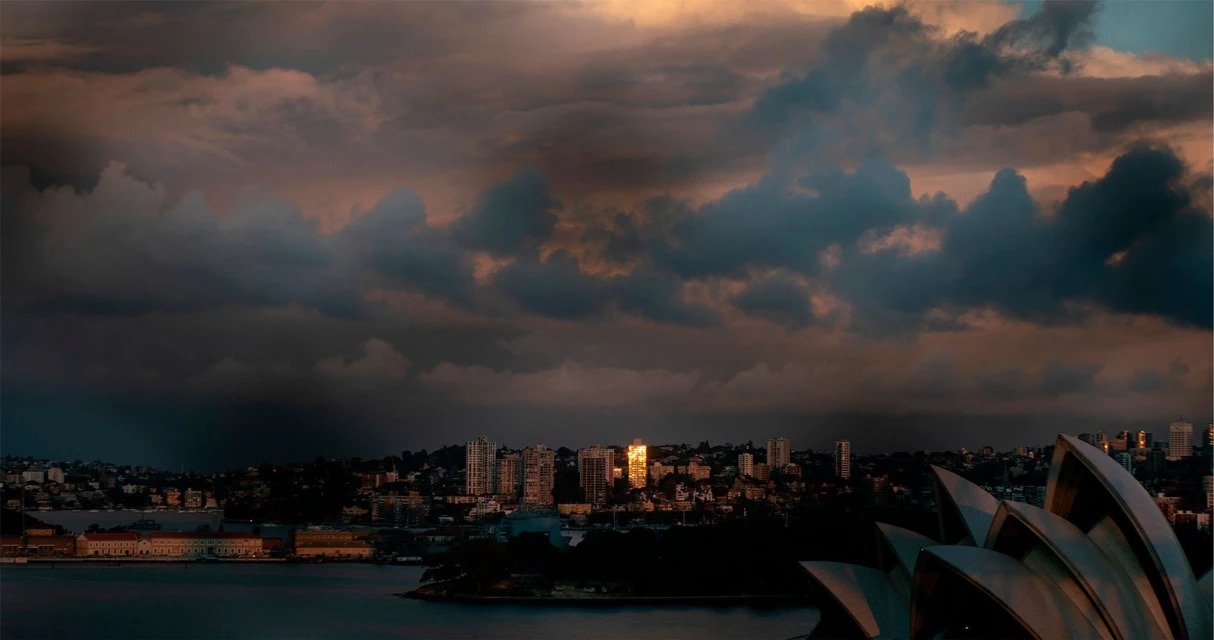 Panoramic cityscape view of the Sydney Opera House and skyline at dusk, featuring dramatic clouds and the waterfront.
