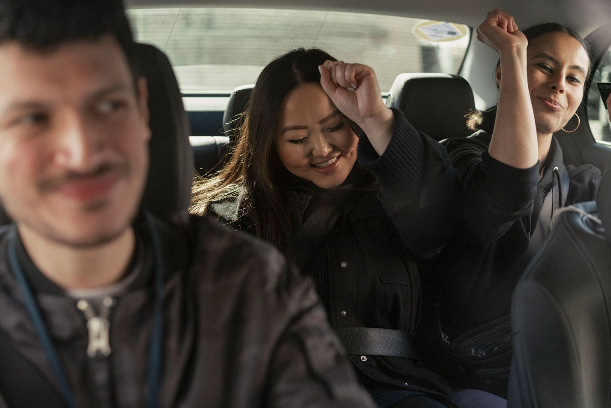 Three smiling people riding in the back seat of a car, two women with raised arms in celebration, and one man blurred in the foreground.