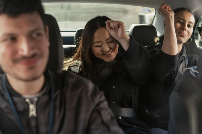 Three smiling people riding in the back seat of a car, two women with raised arms in celebration, and one man blurred in the foreground.