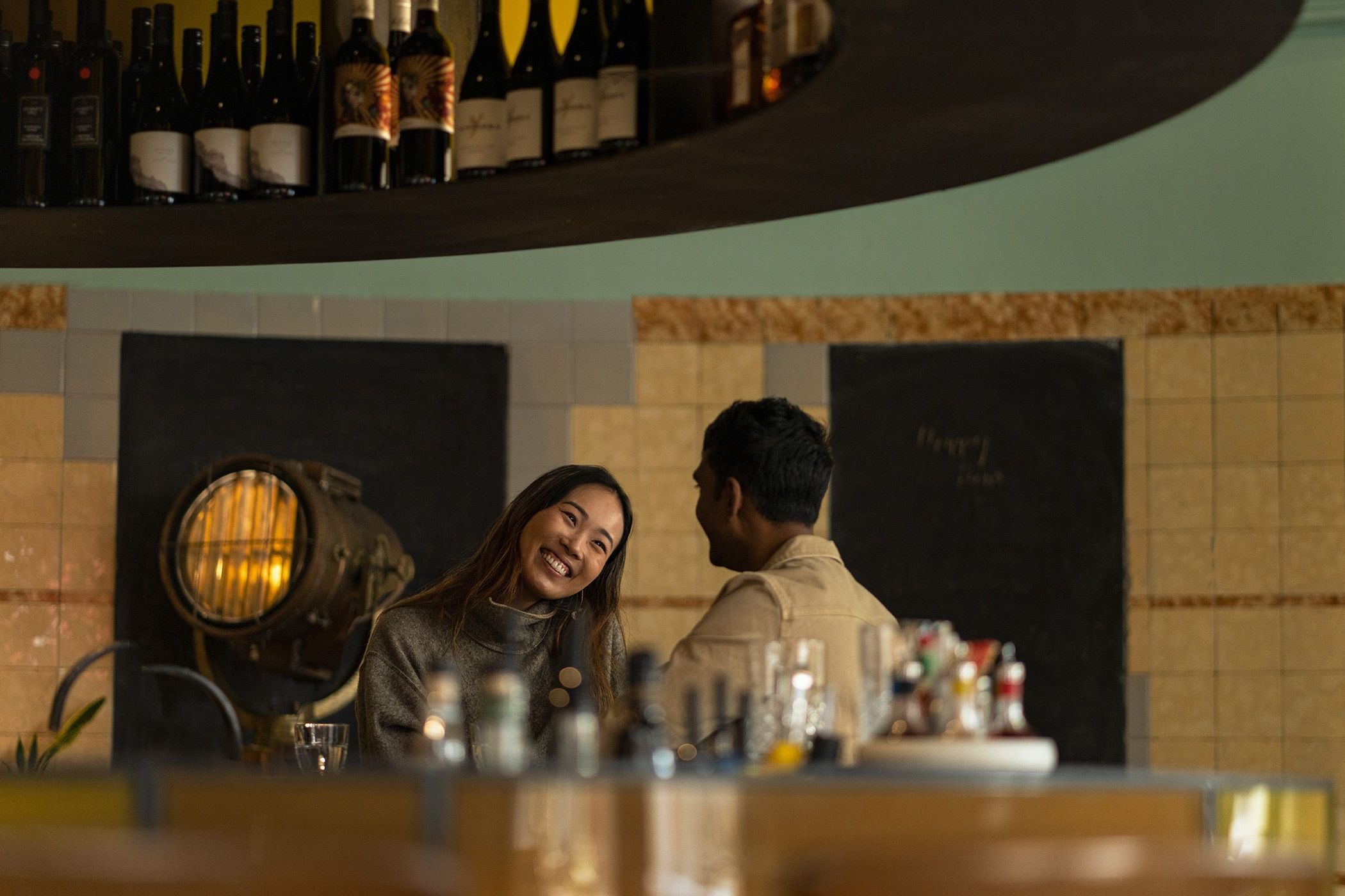 A smiling Asian woman and a man in an indoor restaurant setting, seated at a counter with a display of bottles and glasses.