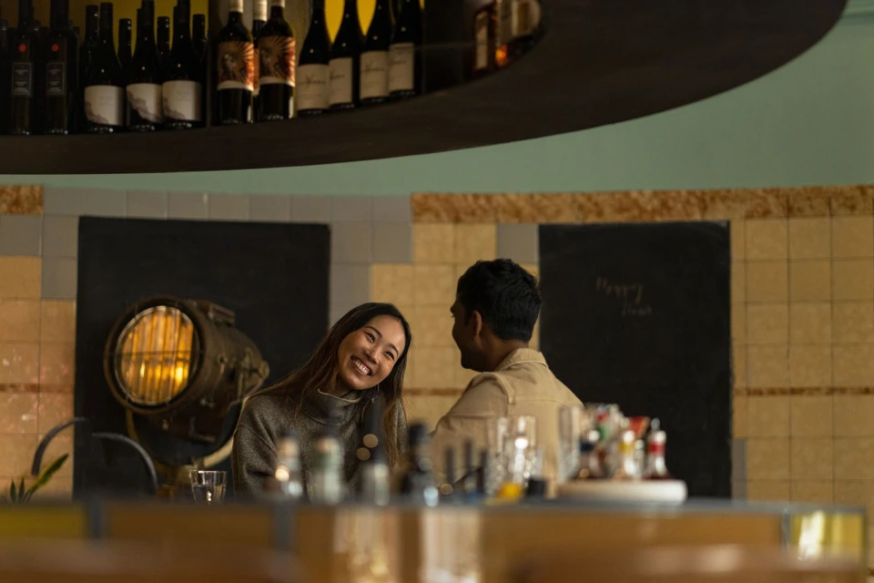 A smiling Asian woman and a man in an indoor restaurant setting, seated at a counter with a display of bottles and glasses.