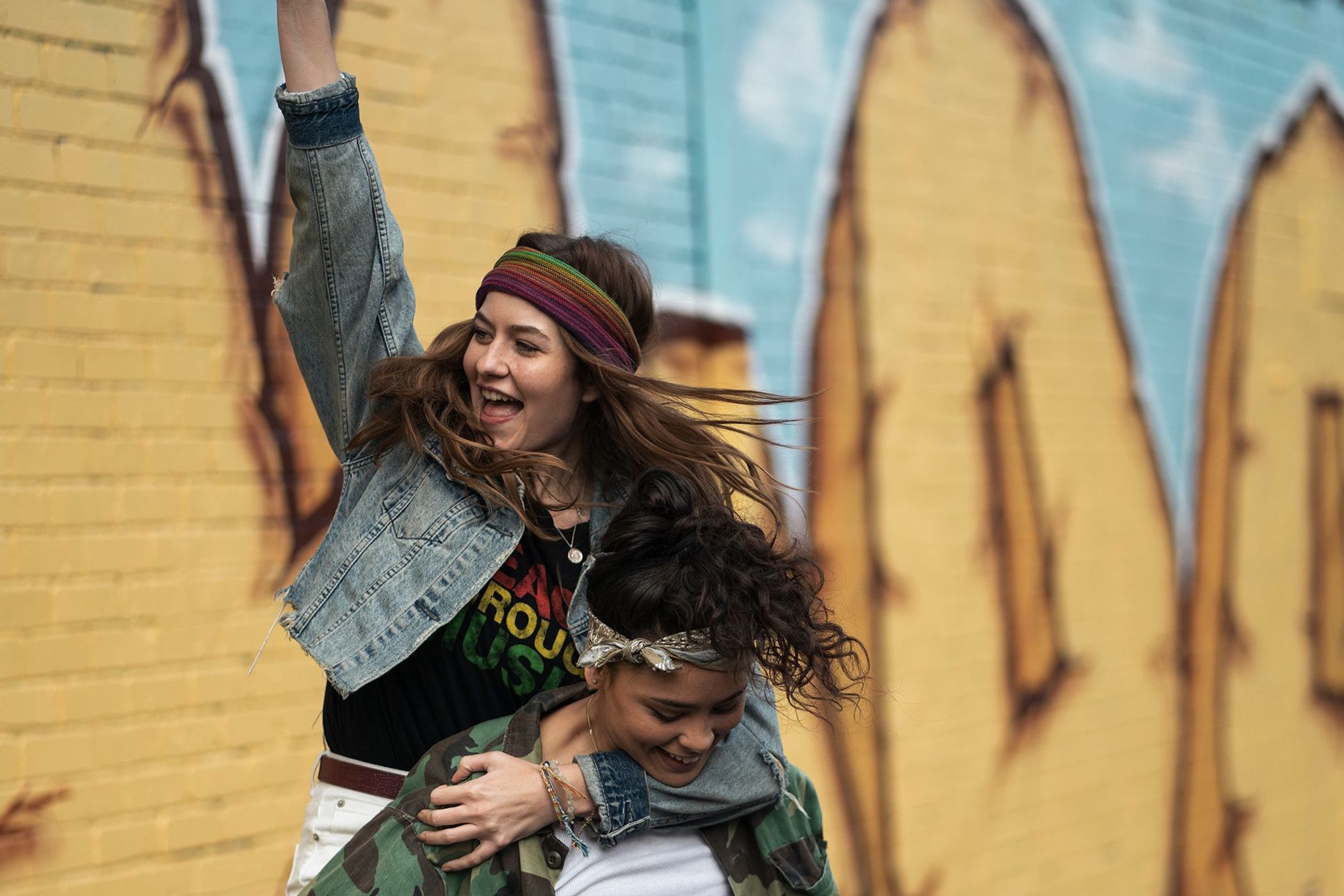 Two young women outdoors: One woman piggybacking while raising her arm in front of a painted wall, showcasing a youthful and carefree scene.