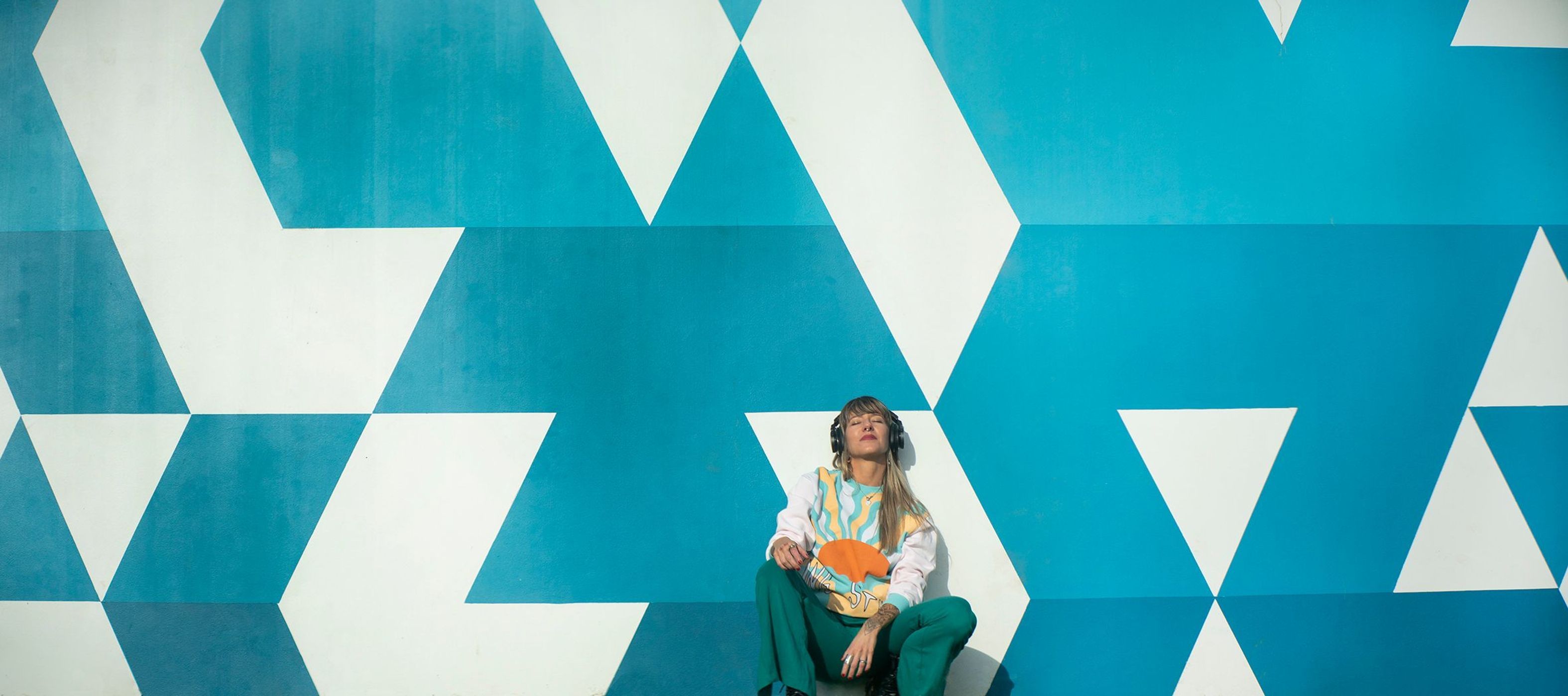 Blonde woman with headphones sits against an aqua and white geometric wall in a long shot, wearing a sweater and matching pants.