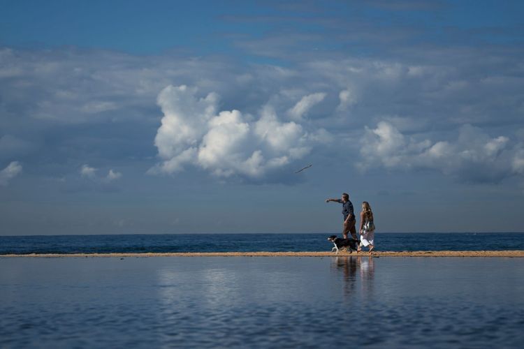 Man and woman walking a dog on a sandy beach with ocean and cloudy sky reflections, enjoying a sunny day outdoors.