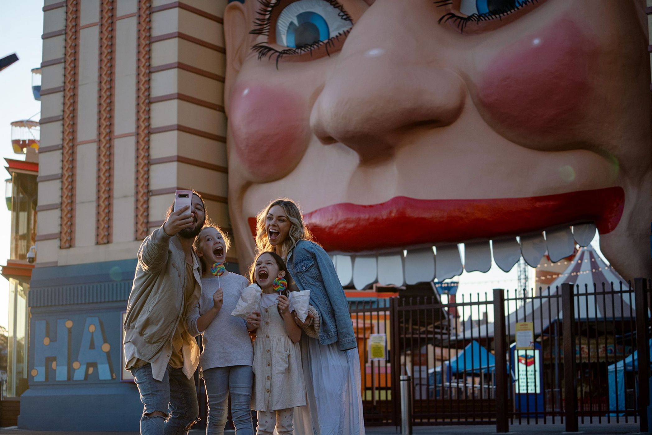Family takes a selfie in front of Luna Park's iconic face entrance in Melbourne, Australia; happy expressions, colorful background.