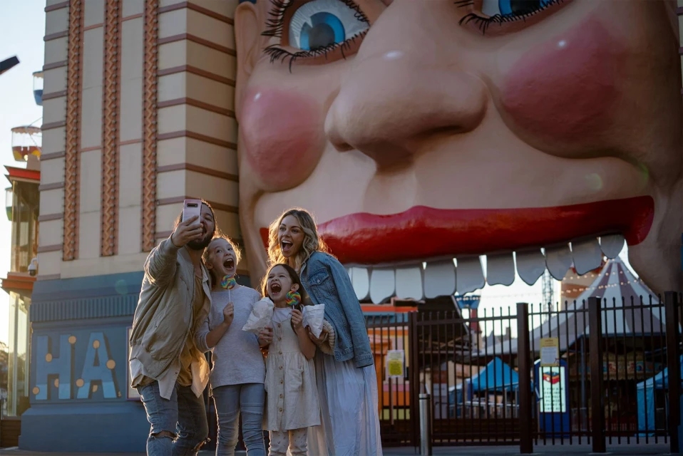 Family takes a selfie in front of Luna Park's iconic face entrance in Melbourne, Australia; happy expressions, colorful background.