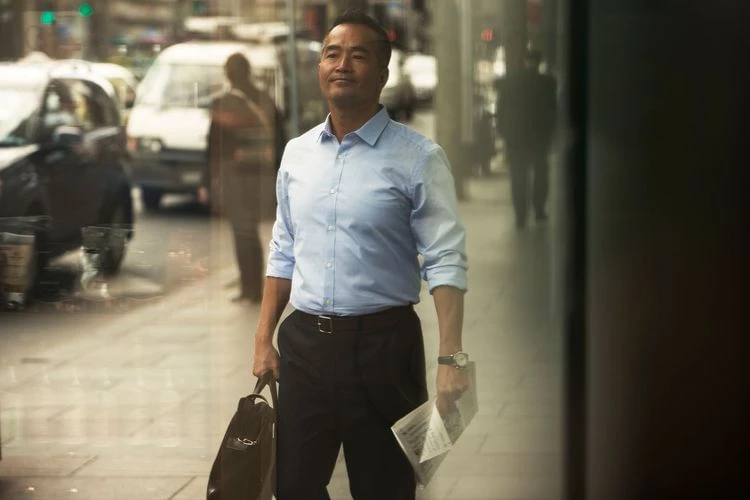 An Asian man in business attire walking outdoors on a city sidewalk, carrying a briefcase and newspaper, with a blurred background.