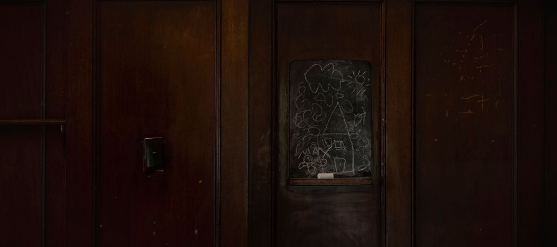 Dark wooden door with a child's chalk drawing of a house, sun, and flowers on a small chalkboard attached to the door's surface.