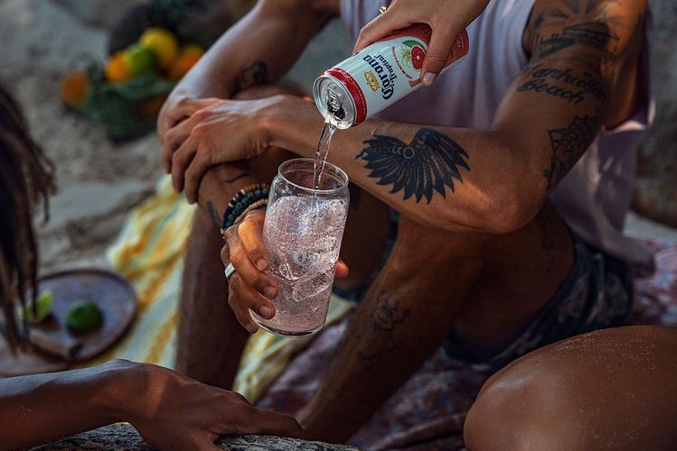 Close-up of an arm pouring Corona Tropical into a glass with ice, set on a beach with a blanket and fruit, suggesting a relaxed outdoor setting.