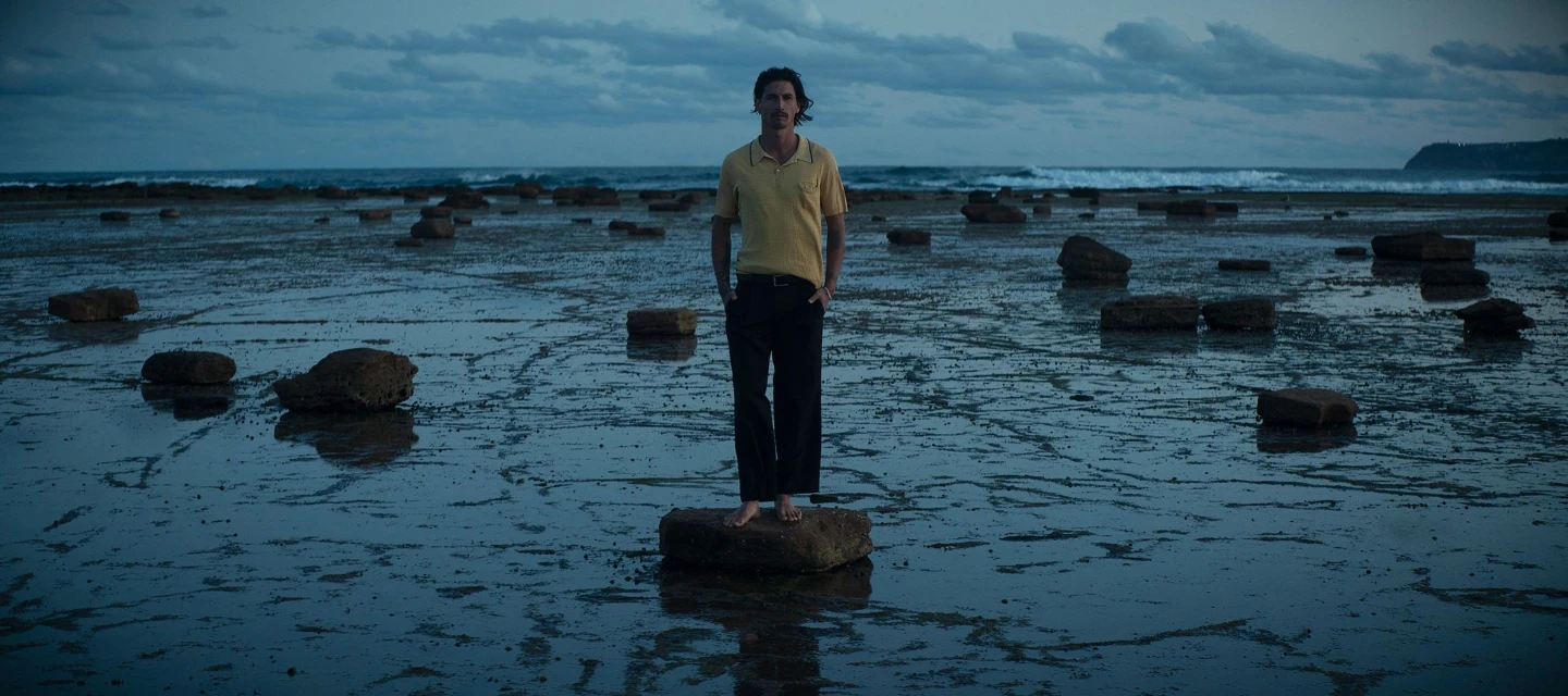 Man in yellow shirt and black pants standing barefoot on rock formation at low tide on a beach at dusk, medium shot.