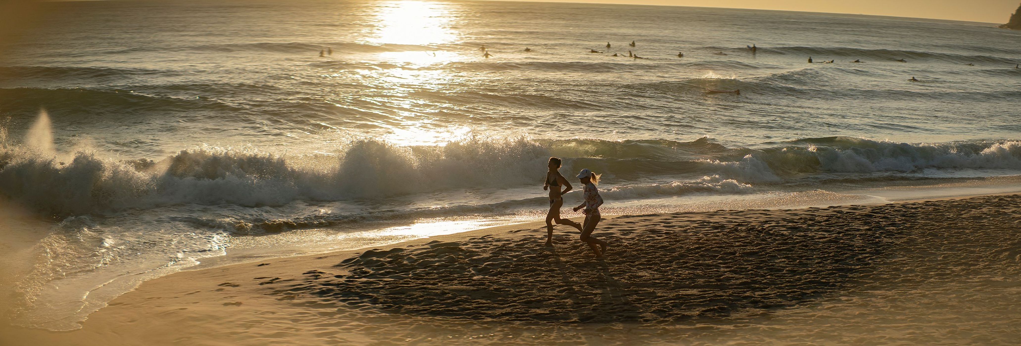Two adult females jog along a sandy beach at sunrise, with gentle ocean waves breaking in the background and surfers in the distance.