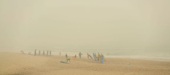 Group of surfers on a sandy beach carrying surfboards under a hazy, smoke-filled sky in Sydney by Dick Sweeney.