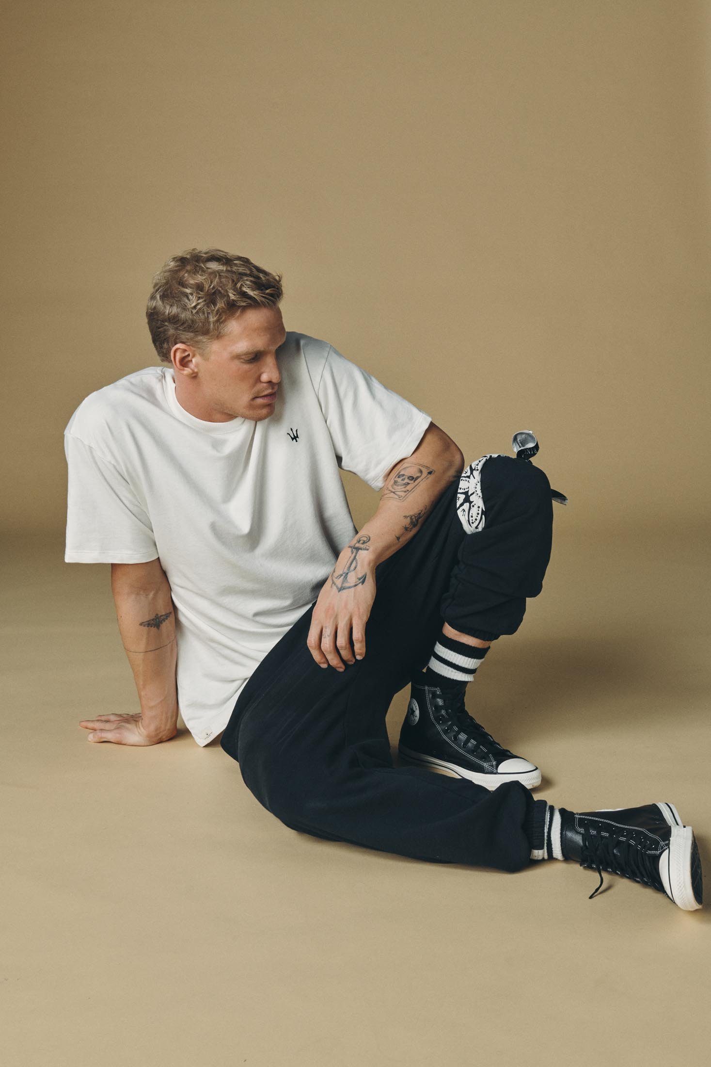 Man in white tee and black sweatpants sitting on tan floor, wearing high-top sneakers and striped socks, posing in a studio setting.