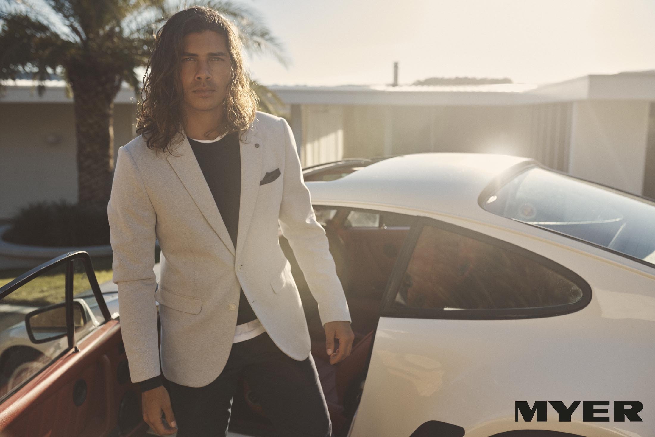 Man in a blazer stands beside a white Porsche, creating a fashionable outdoor automotive shot with palm trees and architecture.