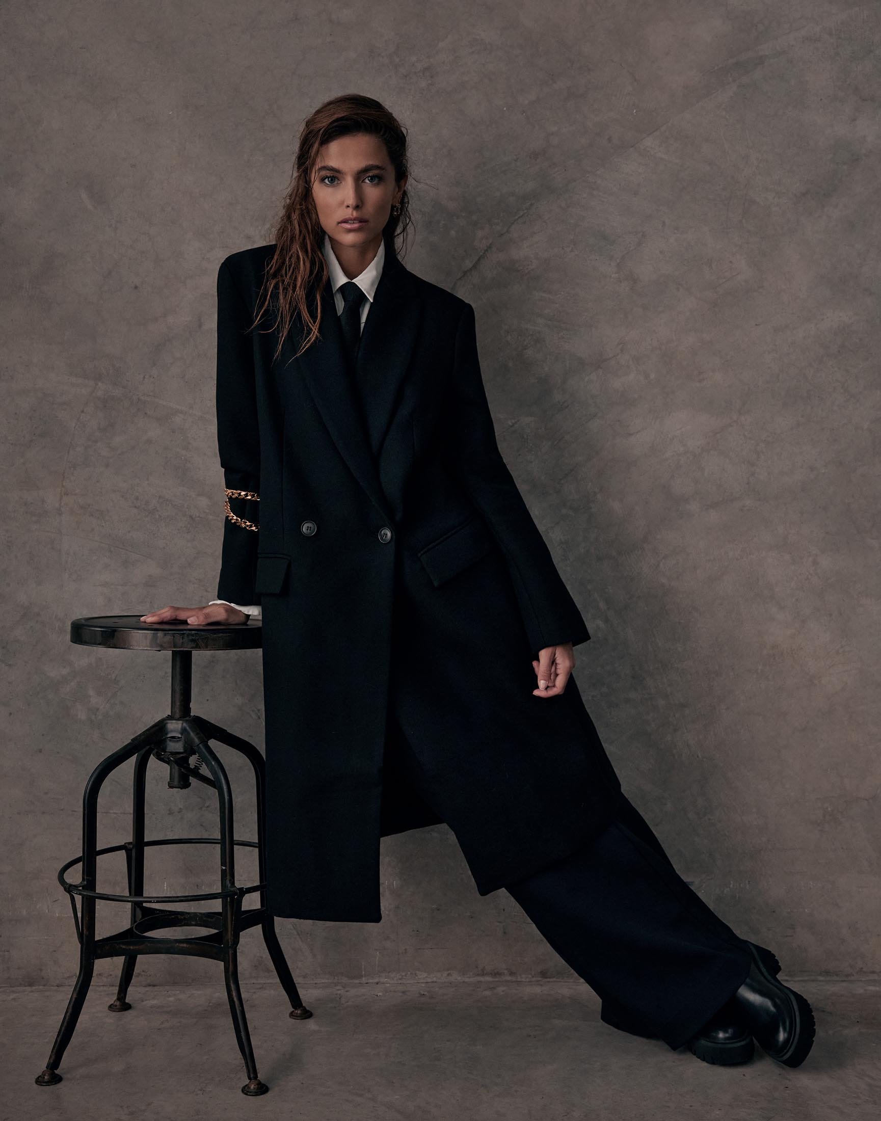 Adult female in a black pantsuit and long coat poses in a studio with a dark backdrop. She leans against an industrial stool.