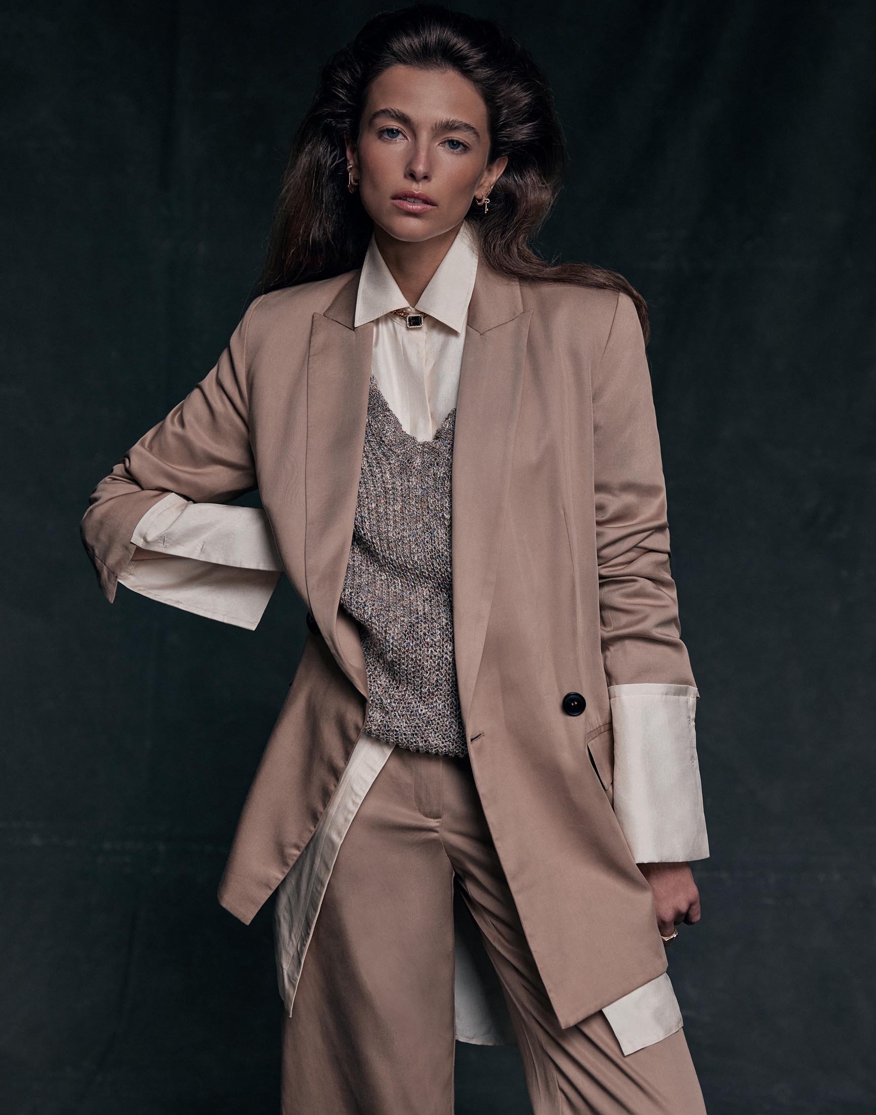 Portrait of a woman wearing a tan blazer and pants over a cream collared shirt and gray sweater vest in a studio setting.