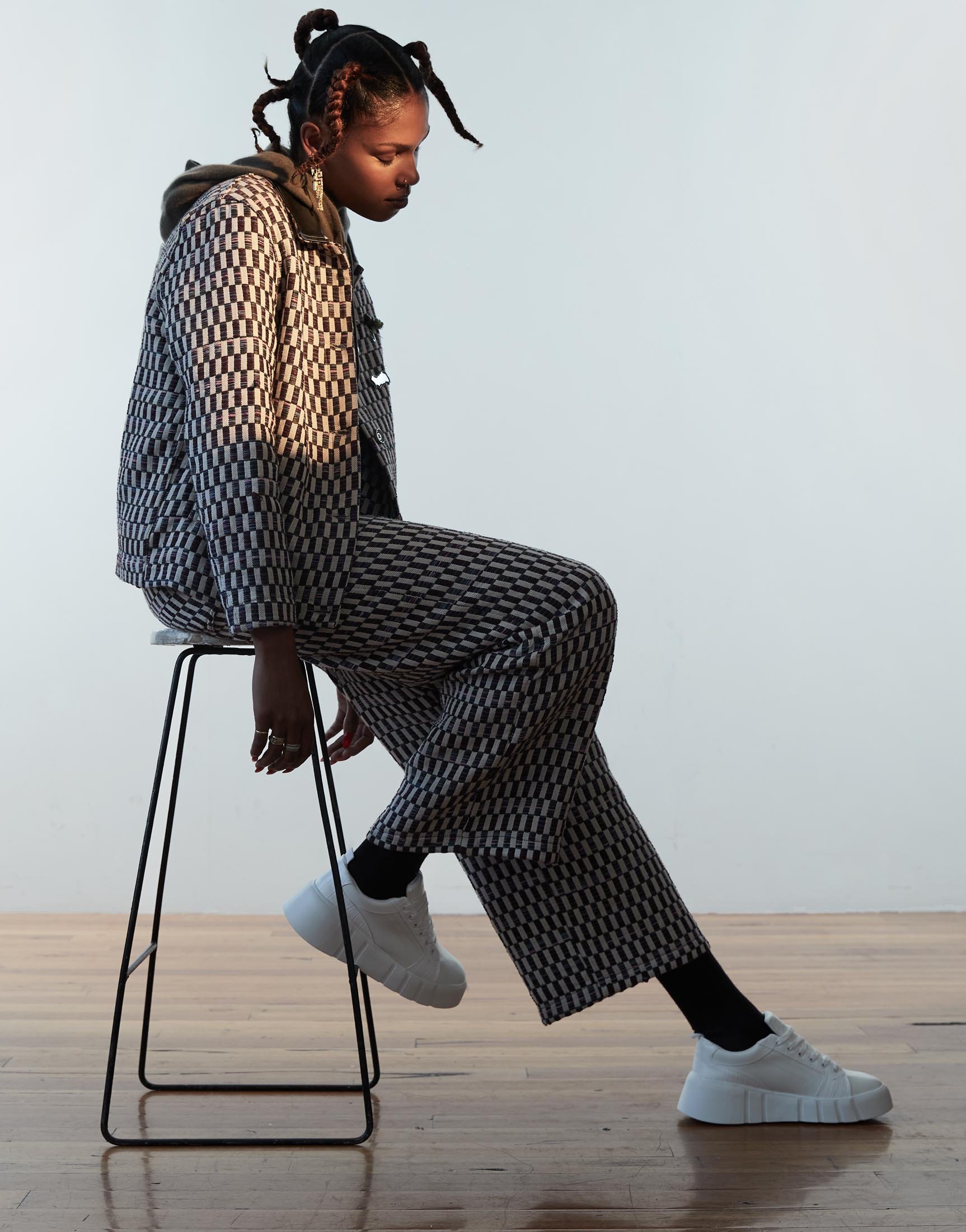 A young woman in a patterned suit sits on a stool in a bright photo studio, looking down; she wears white platform sneakers.