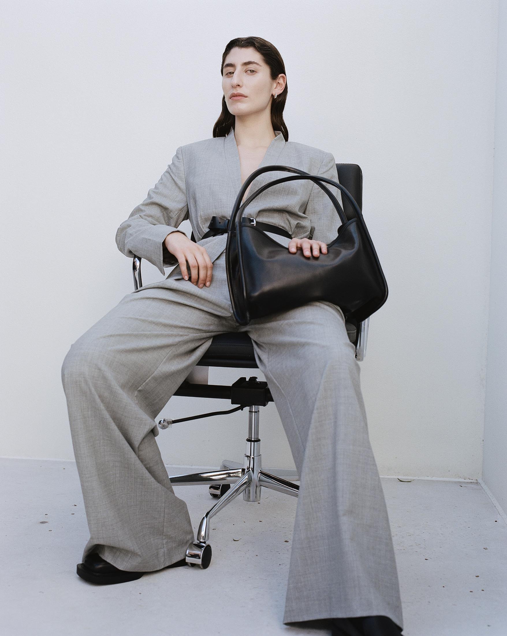 Adult woman in a grey suit sits in a chair, holding a black leather handbag against a white background in a fashion editorial.
