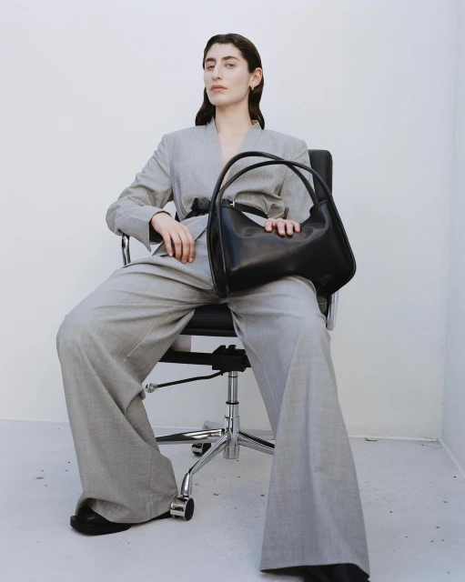 Adult woman in a grey suit sits in a chair, holding a black leather handbag against a white background in a fashion editorial.