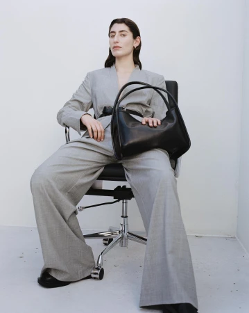 Adult woman in a grey suit sits in a chair, holding a black leather handbag against a white background in a fashion editorial.