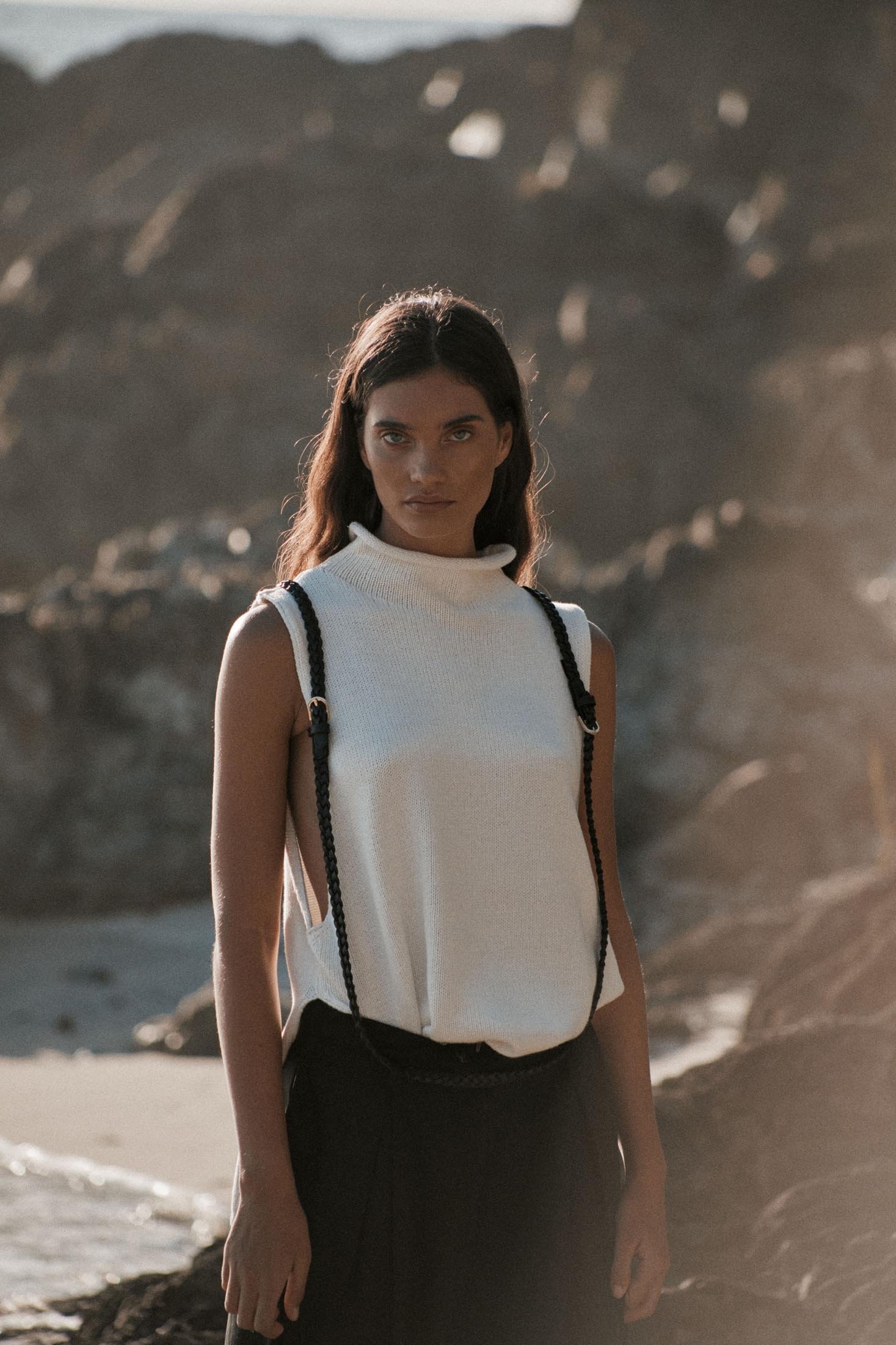 Brunette woman in a white knit top and black pants poses on a rocky beach with ocean and a distant shoreline in the background.