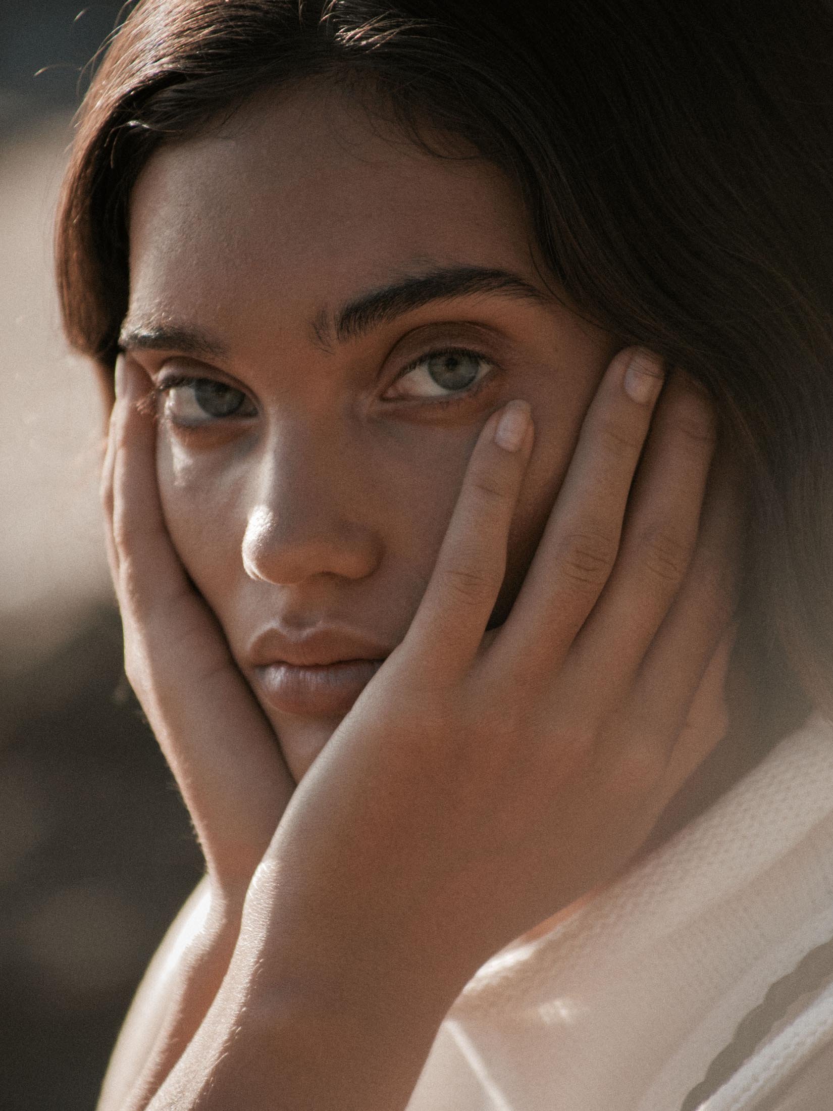 Close-up portrait of a woman with brown hair and green eyes, with her hands framing her face, set against a blurred neutral background.