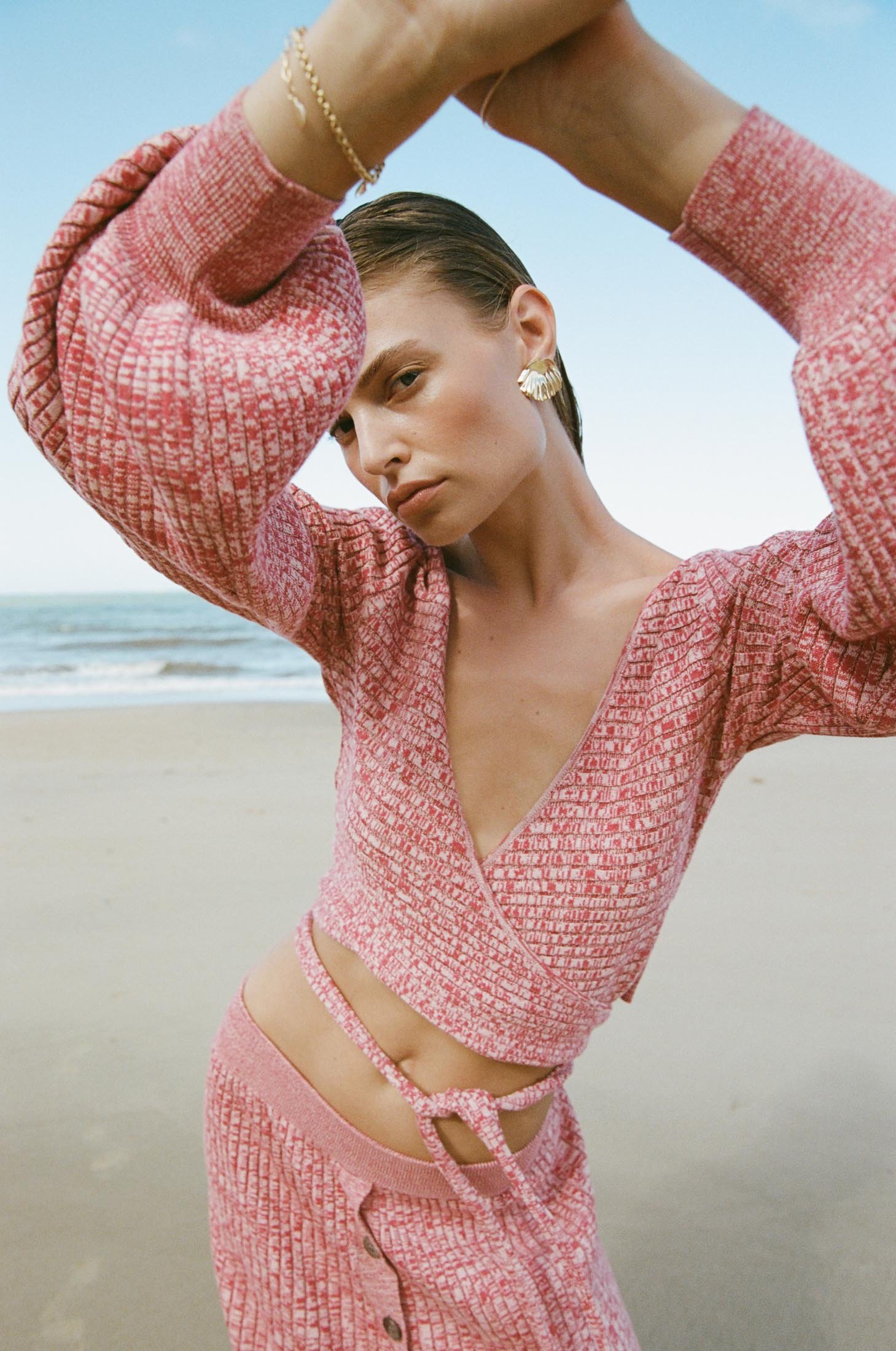 A woman in a pink knit outfit poses outdoors against a beach and sky backdrop, showcasing fashion and beauty in a lifestyle context.