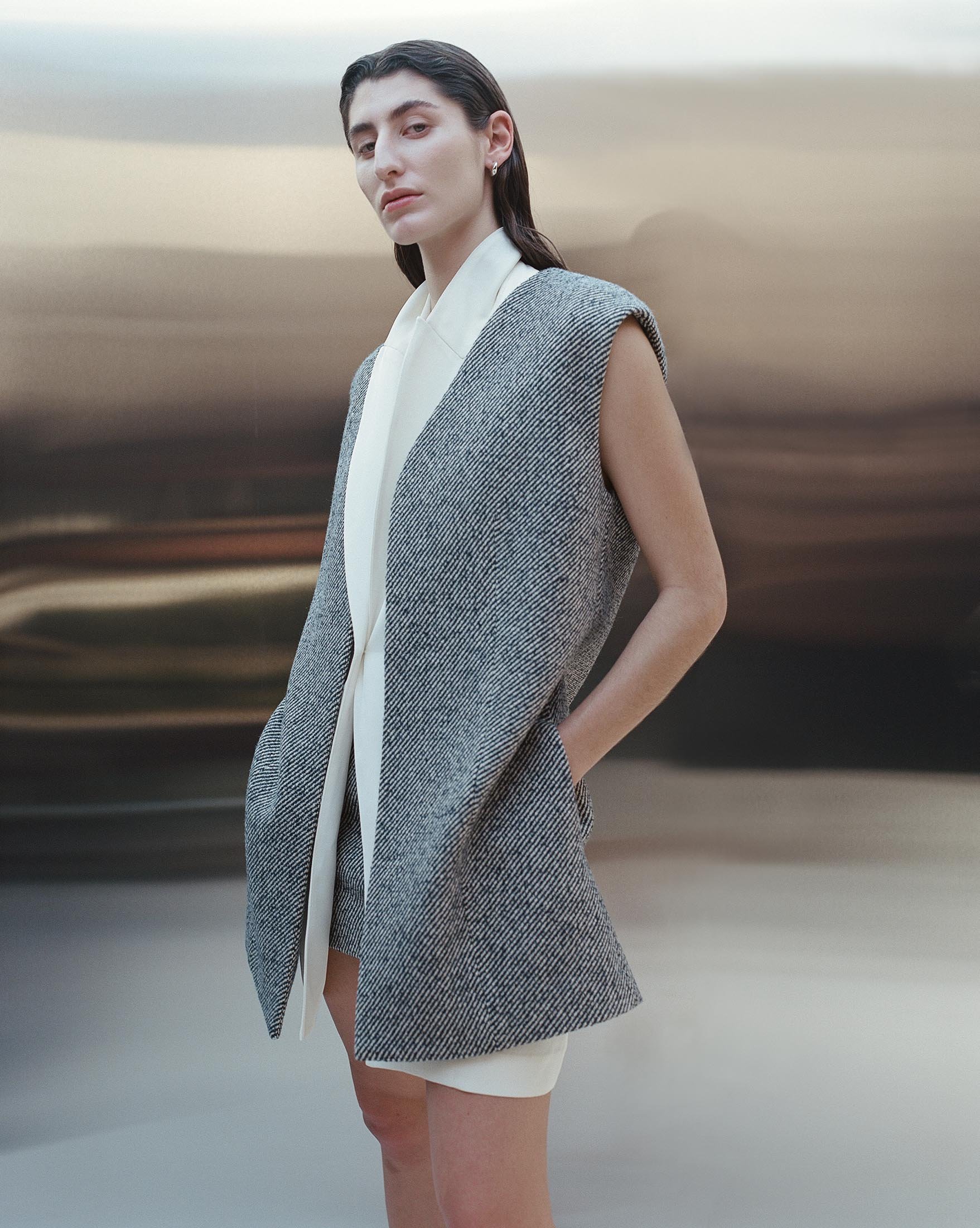 Fashionable woman poses in sleeveless grey tweed blazer and white dress against blurred neutral background in a studio setting.