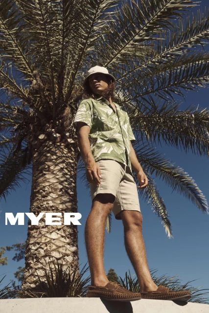 A male model poses outdoors wearing a green leaf-print shirt, khaki shorts, bucket hat, and loafers against a backdrop of a palm tree and blue sky.