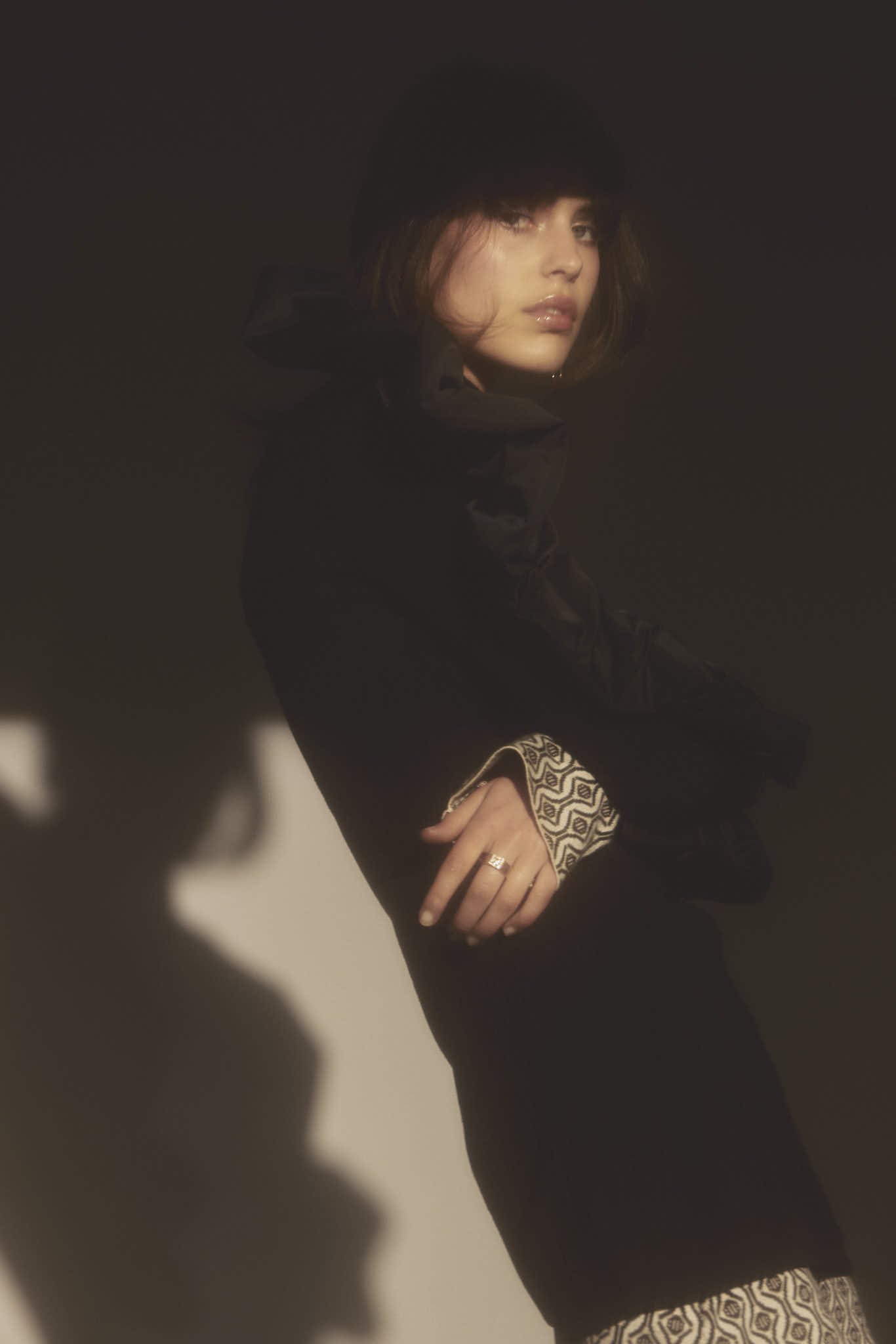 Indoor portrait of a young woman wearing a dark coat and fur hat, with dramatic lighting and a blurred shadow creating a moody atmosphere.