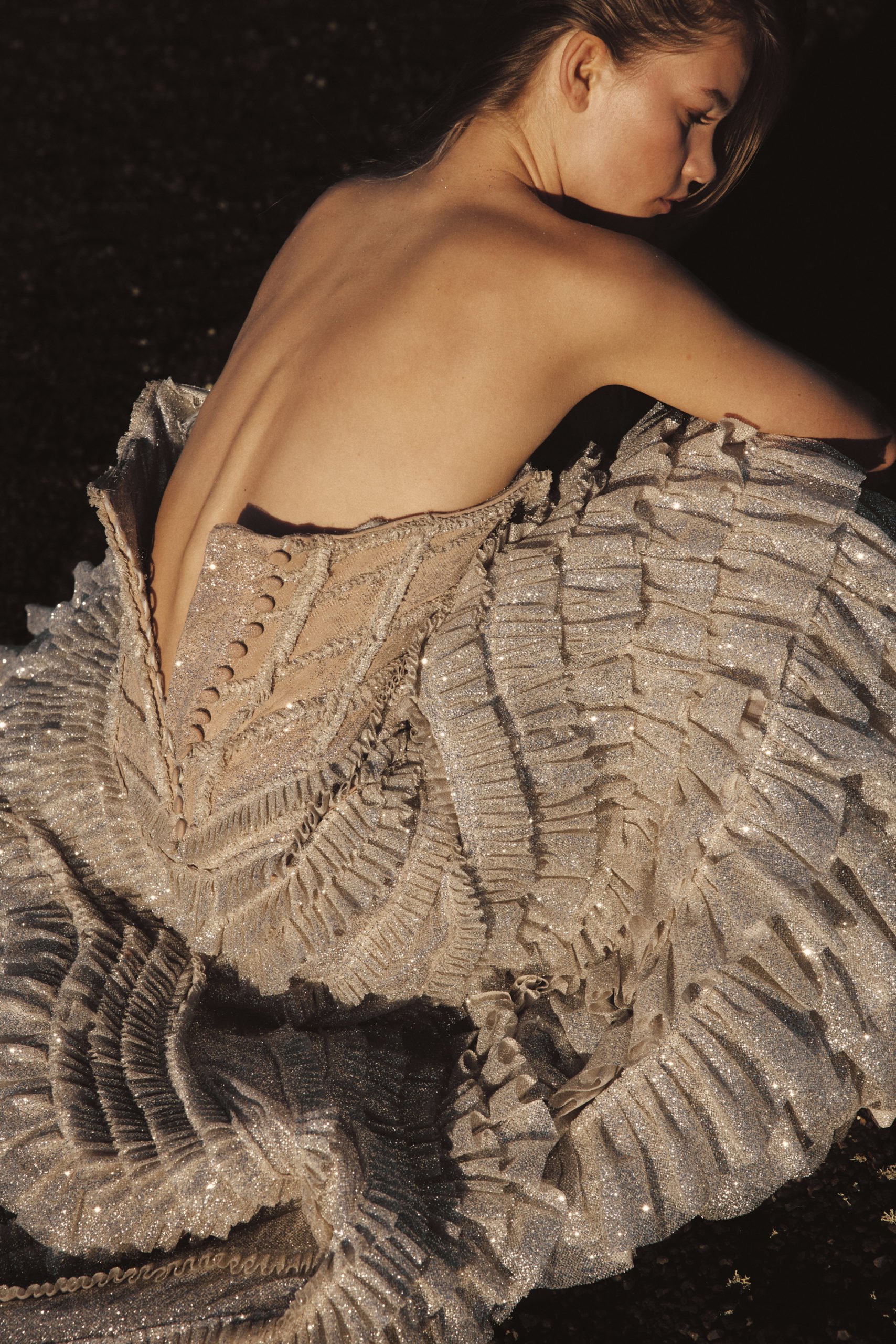 Woman with bare back wearing elaborate shimmering gown with ruffles, photographed in soft light against dark background in portrait orientation.