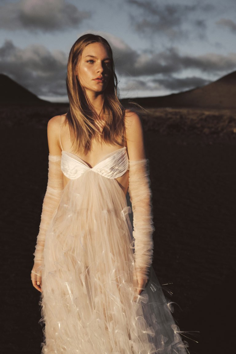 Woman in a sheer white dress and long, tulle sleeves stands outdoors on a rocky landscape with mountains and a cloudy sky in the background.