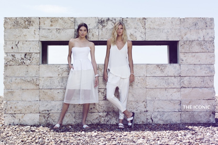 Two women in white outfits pose against a modern stone wall under a bright sky, showcasing fashion and minimalist style.