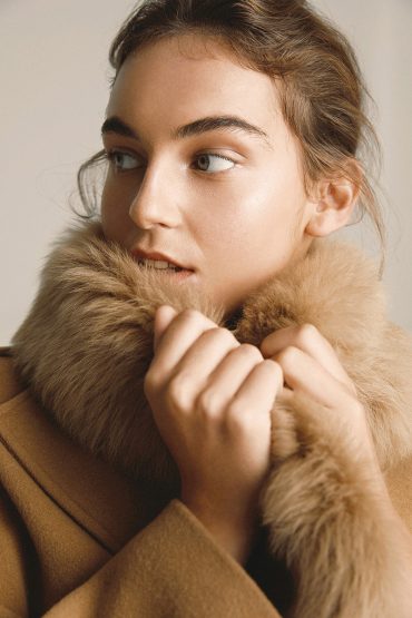 A young woman poses in a medium shot, wearing a tan coat with a large fur collar. The scene is brightly lit in a photo studio.