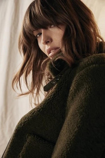 Close-up shot of an adult female model with wavy brunette hair, wearing a fuzzy green jacket, posing in a neutral indoor setting with soft lighting.