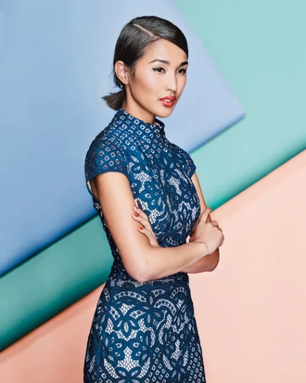 Portrait of an Asian woman wearing a navy blue lace dress with her arms crossed, standing against a colorful geometric background.