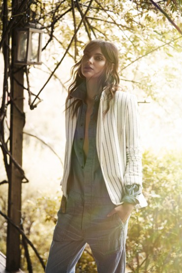 Adult female fashion model wears a white pinstripe jacket and blue jumpsuit, posing outdoors near a weathered lantern.
