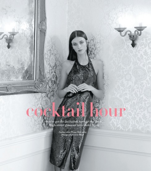 Elegant brunette woman poses in a shimmering grey cocktail dress indoors with a floral wallpaper background, beneath a sconce.