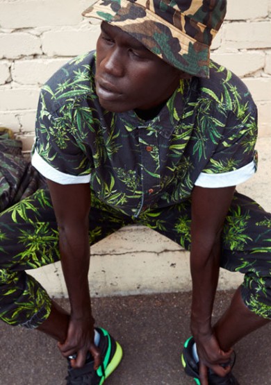 Man poses in patterned shirt and pants with bucket hat against brick wall, looking stylish in urban setting, with black and neon sneakers.
