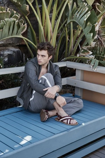 Adult male sitting in a cross-legged pose on a blue wooden platform with tropical plants in the background, wearing sandals and casual clothing.