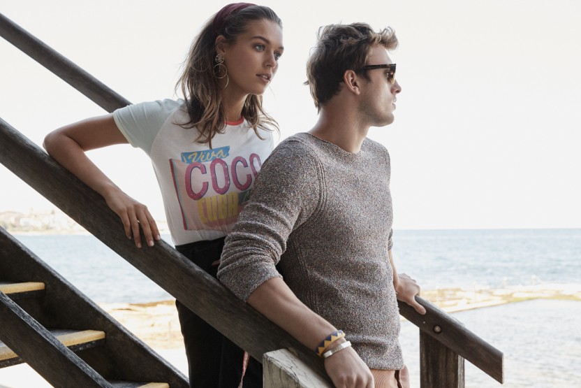 Outdoor fashion portrait of a young couple on stairs overlooking the sea. The woman wears a t-shirt; the man, a sweater and sunglasses.