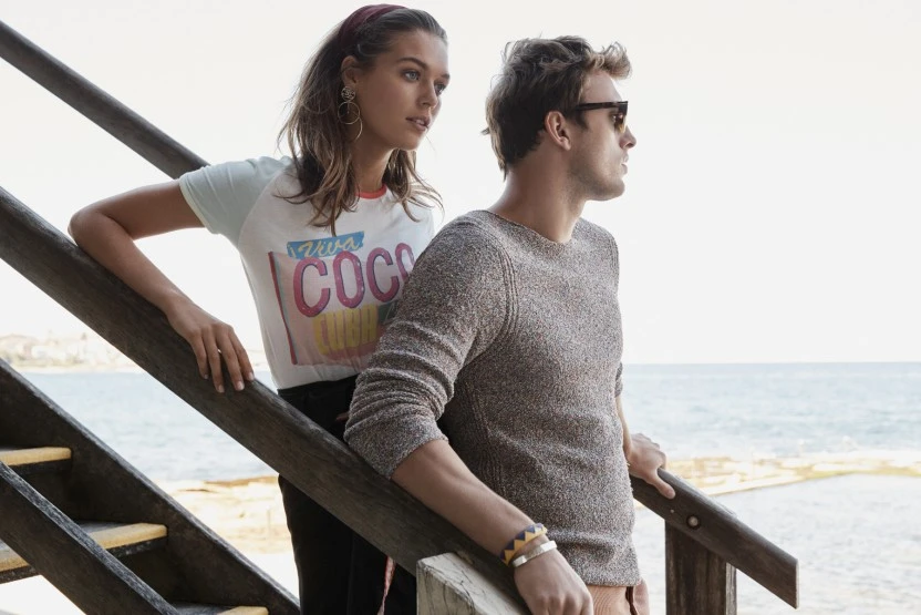 Outdoor fashion portrait of a young couple on stairs overlooking the sea. The woman wears a t-shirt; the man, a sweater and sunglasses.
