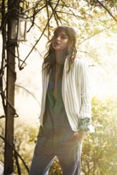 A woman wearing a striped blazer and denim jumpsuit stands outdoors beneath tree branches with sunlight filtering through.