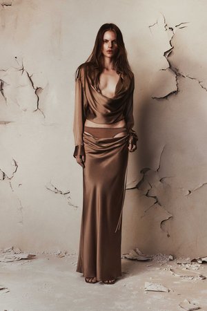 A full-length portrait of a fashion model in a brown silk top and long skirt against a distressed wall, capturing an editorial aesthetic.