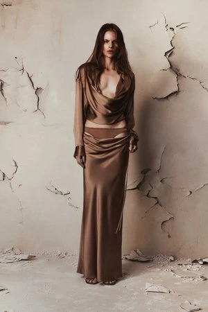 A full-length portrait of a fashion model in a brown silk top and long skirt against a distressed wall, capturing an editorial aesthetic.